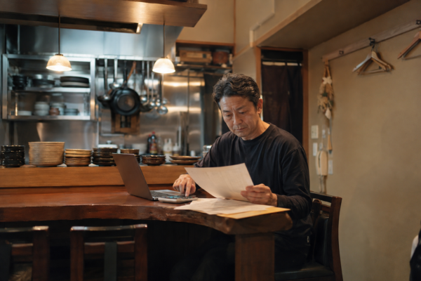 営業後の小規模飲食店で、請求書と電卓、パソコンを見ながら経営数字を確認する店主